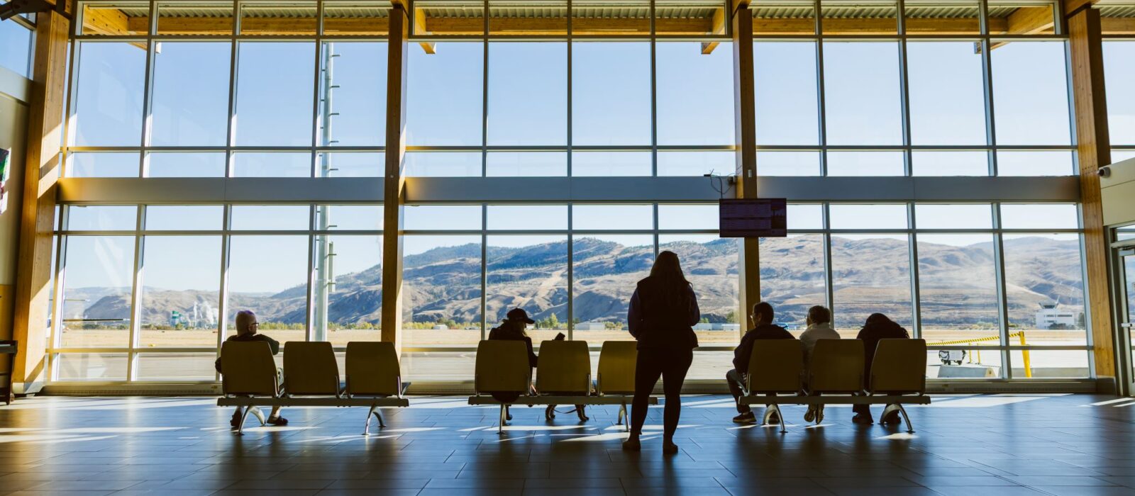 An interior view of the Kamloops Airport arrivals hall, looking outside towards the opposing mountains. A few people are seen waiting for an arriving flight, silhouetted against the windows.